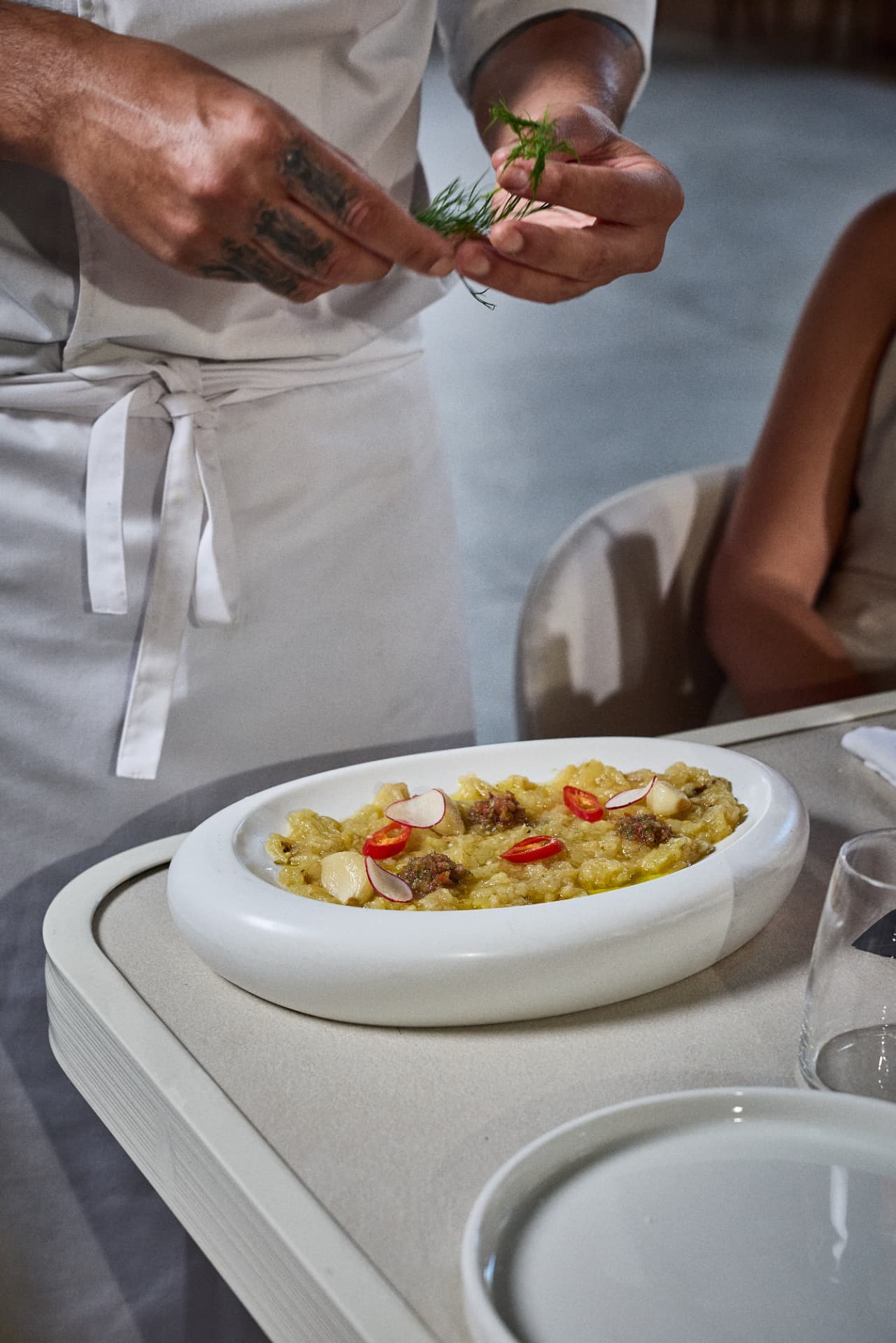 ידי שף מפזרות שמיר על מנת חציל קלוי עם צנונית וצ׳ילי בצלחת לבנה סגלגלה